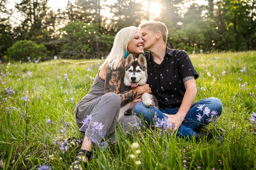 Two women kiss while they hold their dog in a field of flowers