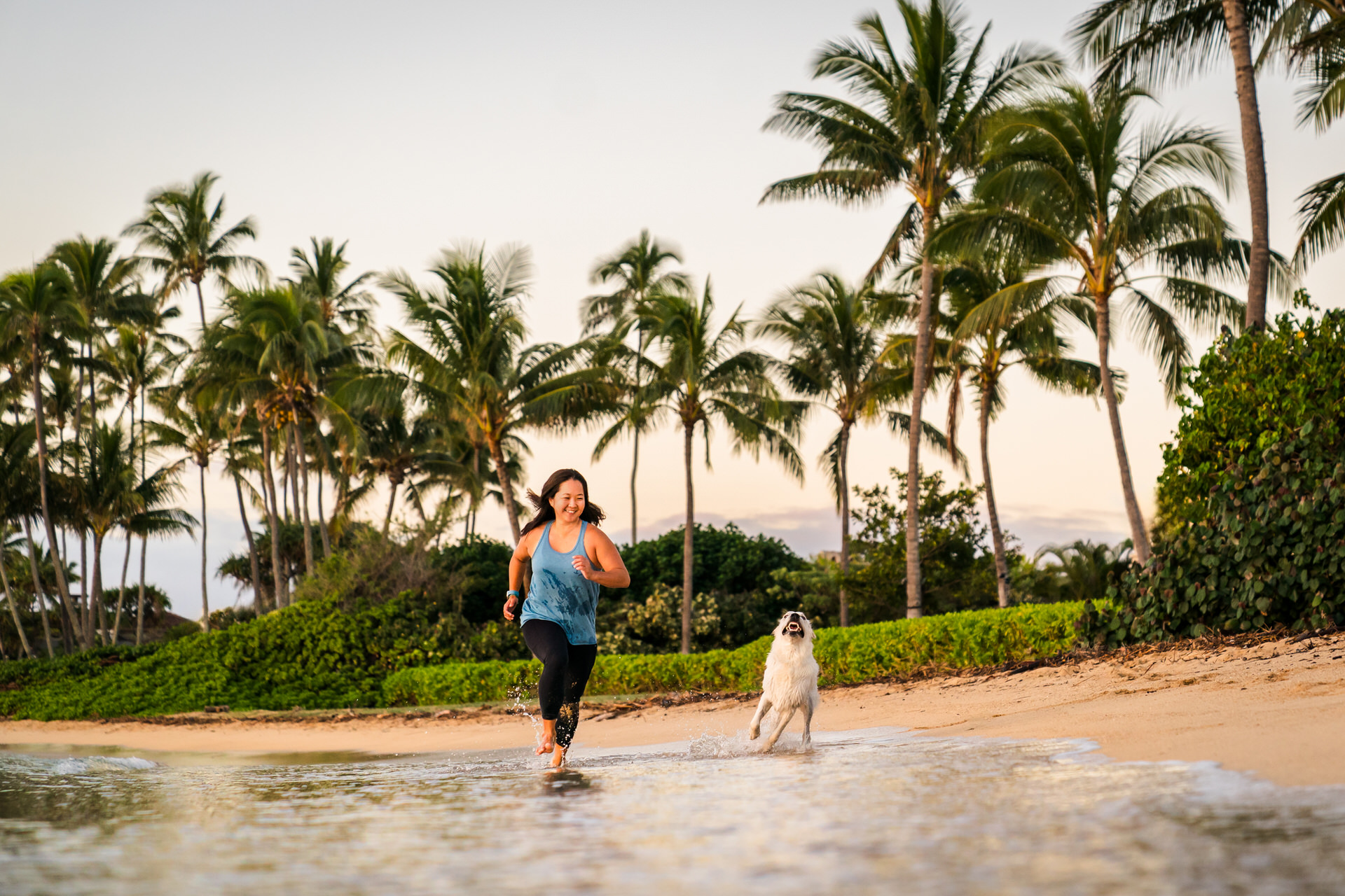 Woman and dog running on tropical beach at sunset